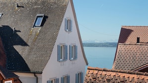 Un edificio con techo inclinado, varias ventanas con contraventanas y vista a un cuerpo de agua y montañas a lo lejos.