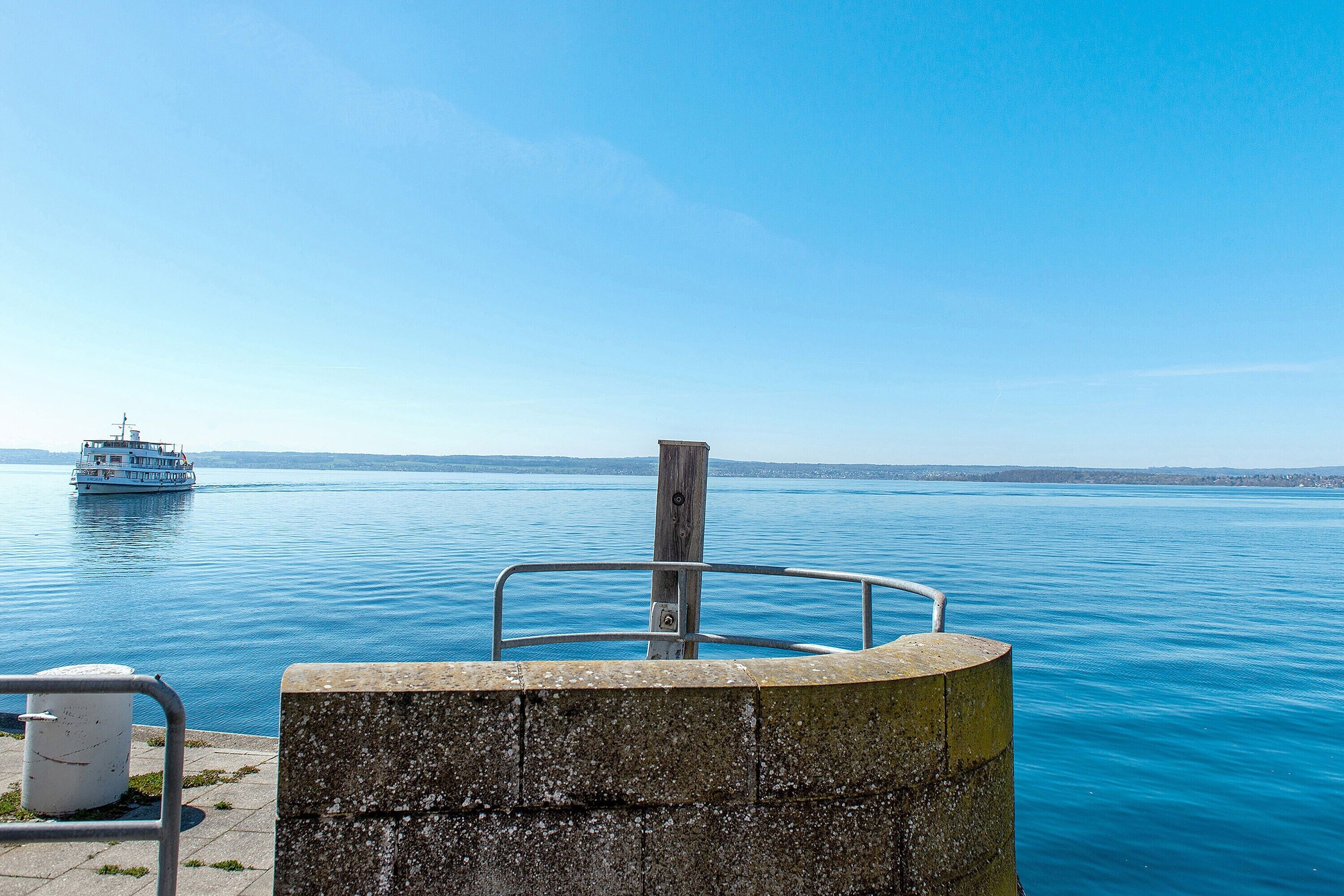 Une vue sur le bord de l’eau avec un bateau sur l’eau, une structure en béton dotée d’une rambarde et un mur de pierre.