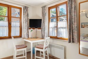 A room with a wooden desk, two white chairs, a television, and large windows with curtains.