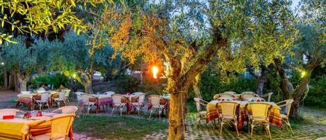 An outdoor dining area with tables and chairs under trees, surrounded by greenery.