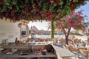 Zona pranzo all'aperto con angolo cottura, sedie e vista sugli edifici sullo sfondo.