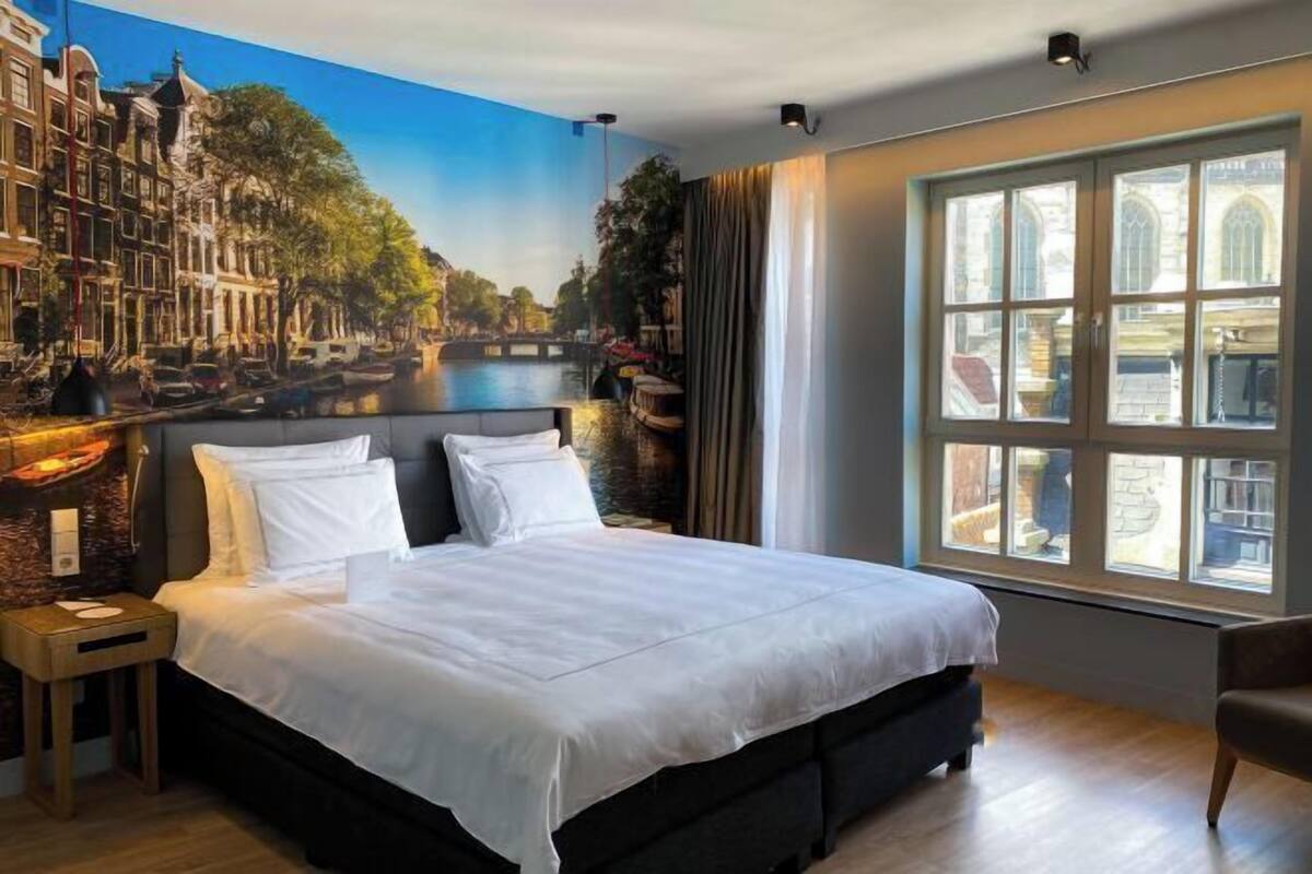 A hotel room with a large bed, a nightstand, a chair, a window with a view of buildings, and a mural of a canal scene.