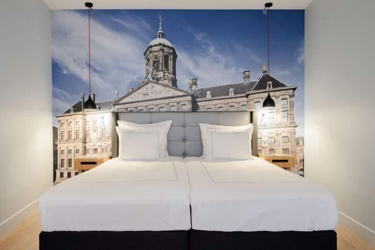 A bed with a white headboard and black skirted legs, set against a backdrop of a cityscape with a prominent building and a clear sky.