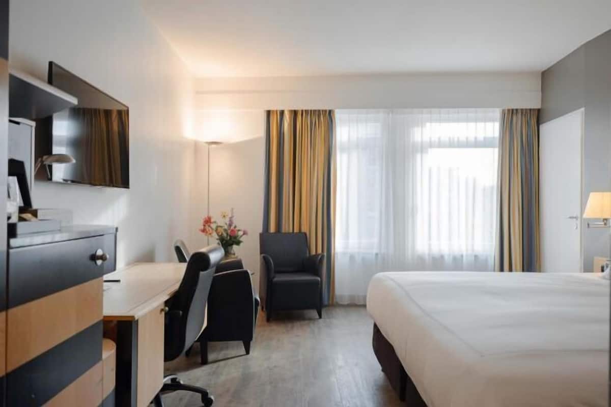 A hotel room with a bed, a desk with a chair, a TV, and a lamp.