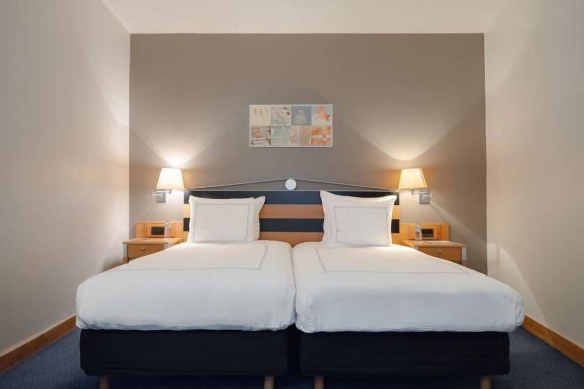 A double bed with white bedding, two bedside tables with lamps, a wall-mounted artwork, and a wooden headboard.