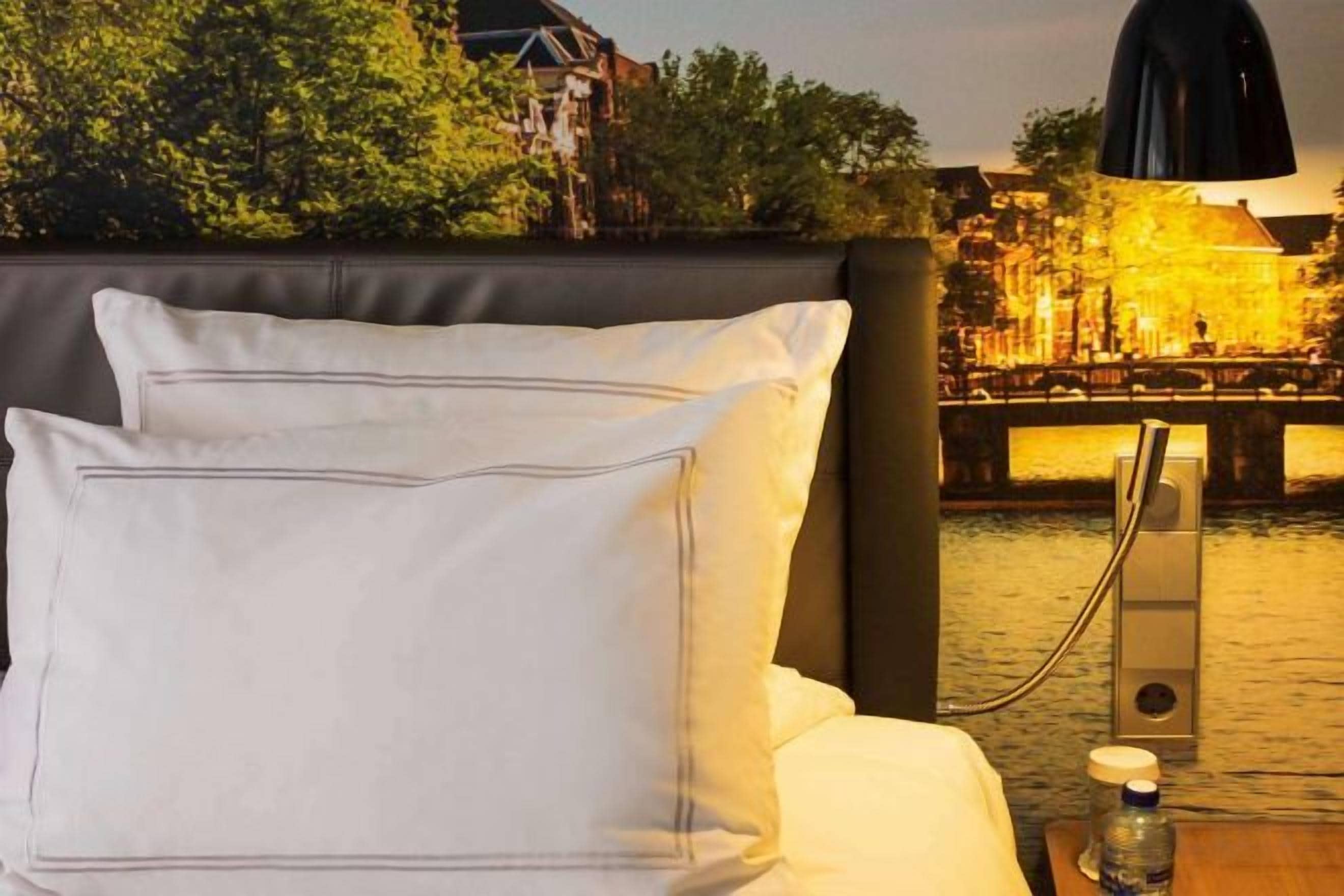 Een bed met witte lakens en een lamp, een fles water, en een uitzicht op een verlichte brug over het water.