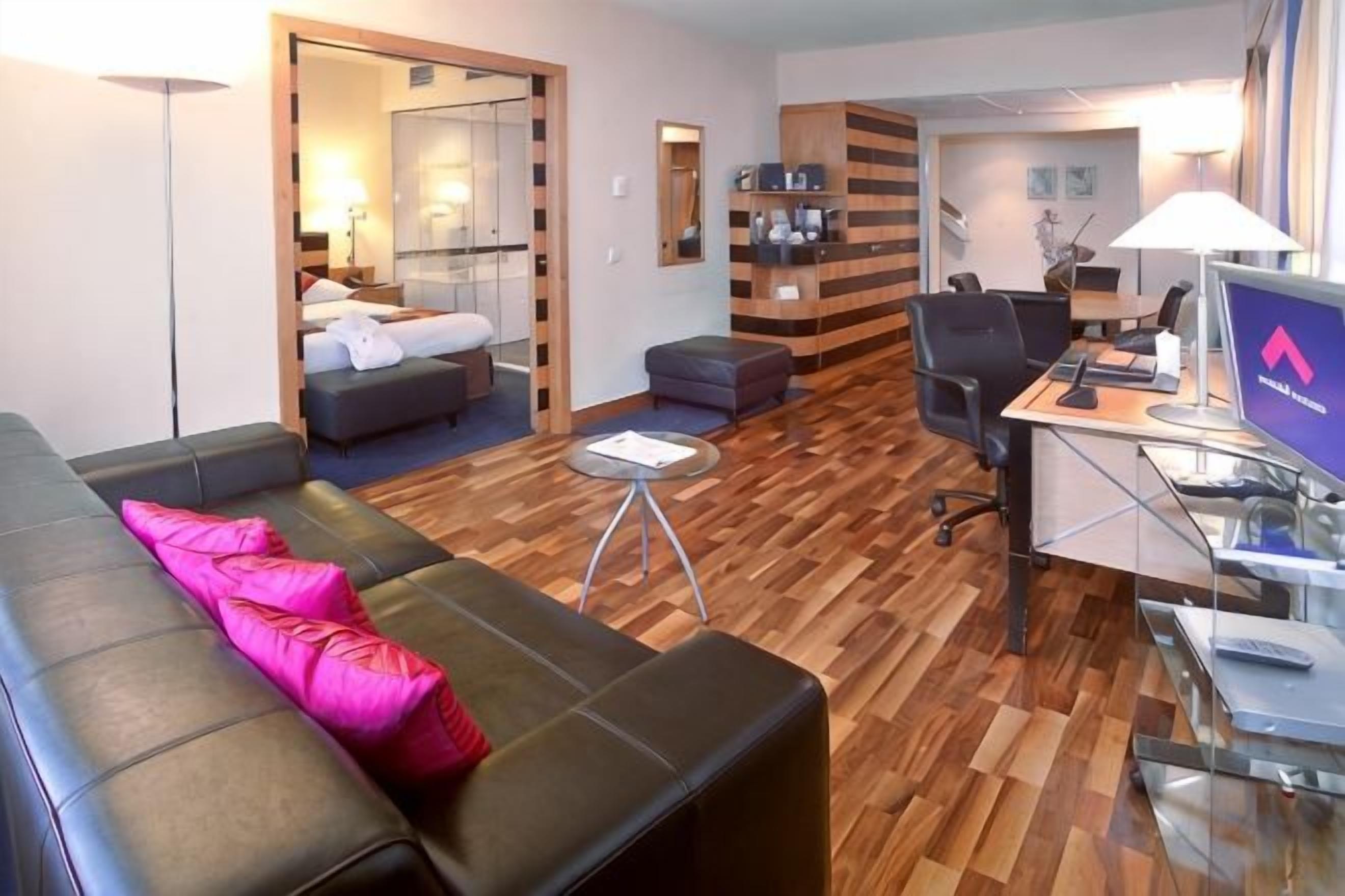 Een moderne hotelkamer met een sofa, een bureau met een computer, een klein tafeltje en een bed met nachtkastje.