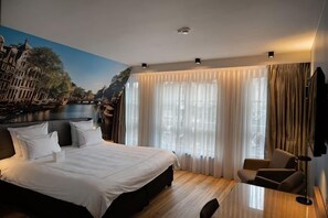 A hotel room with a large bed, a desk, and a view of a cityscape through sheer curtains. - Swissôtel Amsterdam (Amsterdam)
