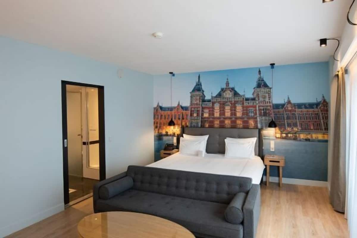 A hotel room with a bed, a grey sofa, a small table, and a mural of a cityscape on the wall.