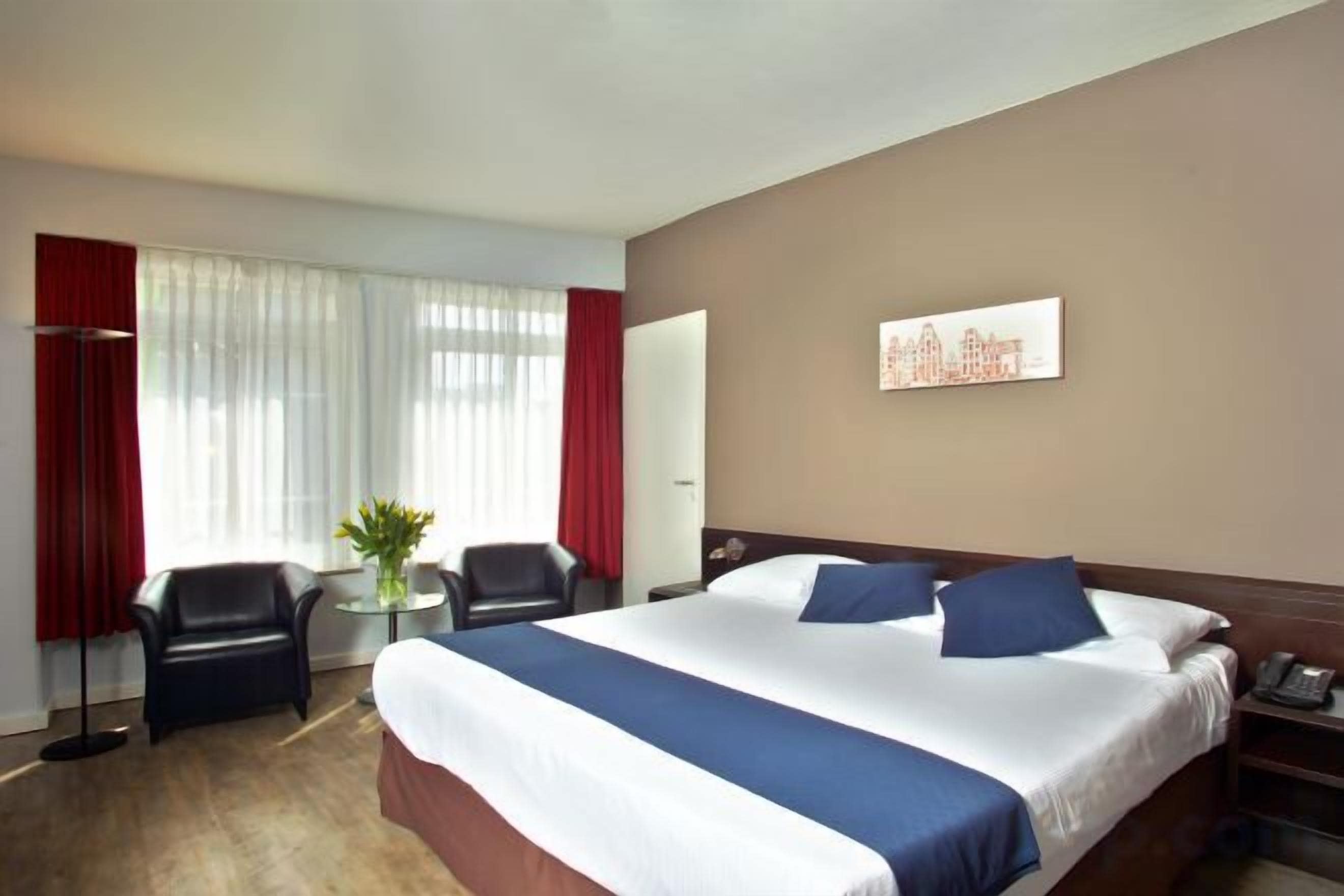 Een hotelkamer met een groot bed, twee stoelen, een bureau en een raam met gordijnen.