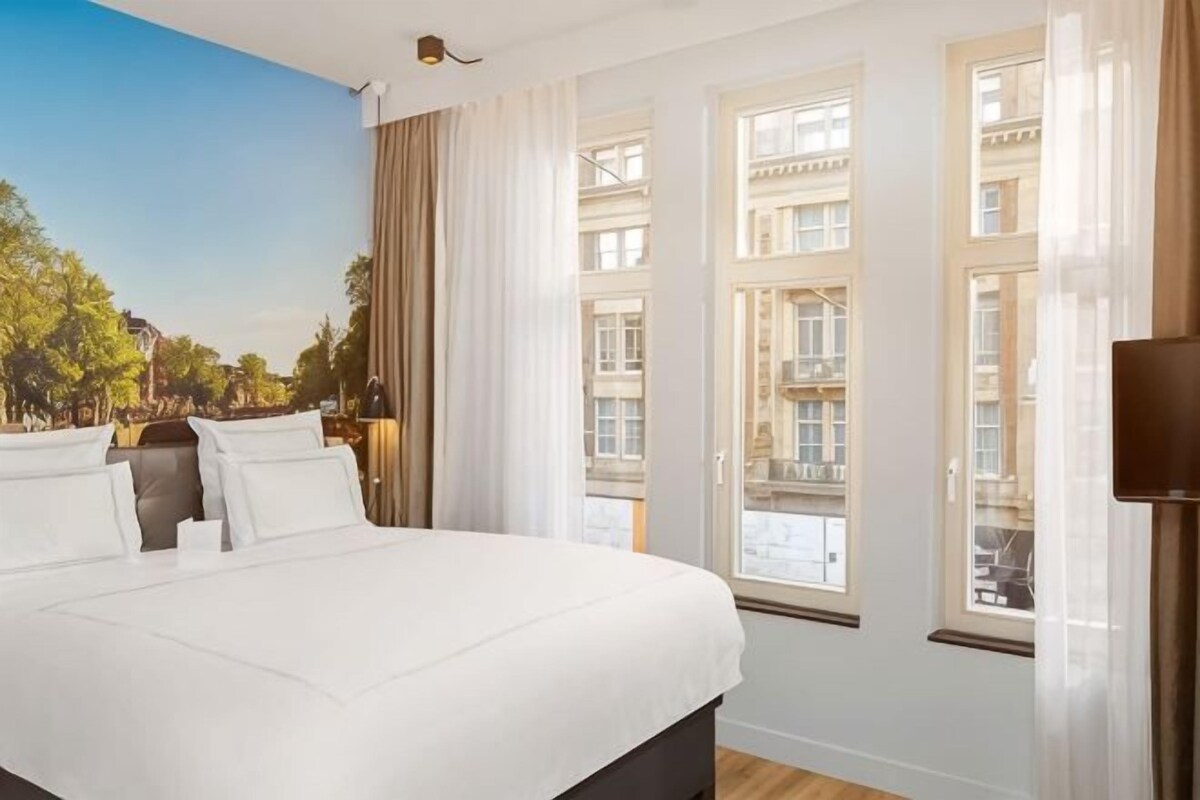 Una habitación de hotel con una cama grande, varias ventanas y vistas a edificios.