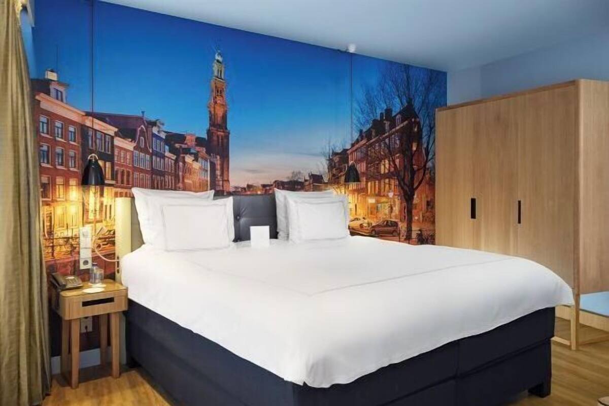 A hotel room with a large bed, a nightstand, a wooden wardrobe, and a cityscape mural on the wall.