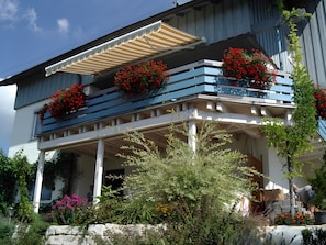 A two-story building with a balcony adorned with red flowers, a covered porch, and a stone wall in front.