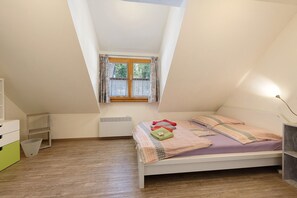 Une chambre avec un plafond en pente, une fenêtre avec des rideaux, un lit recouvert d’une couverture violette et une commode blanche.