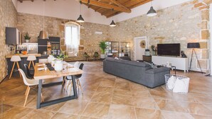 Un espacio habitable amplio con pared de piedra, techo de madera, un sofá gris, una mesa de comedor con sillas y un televisor.