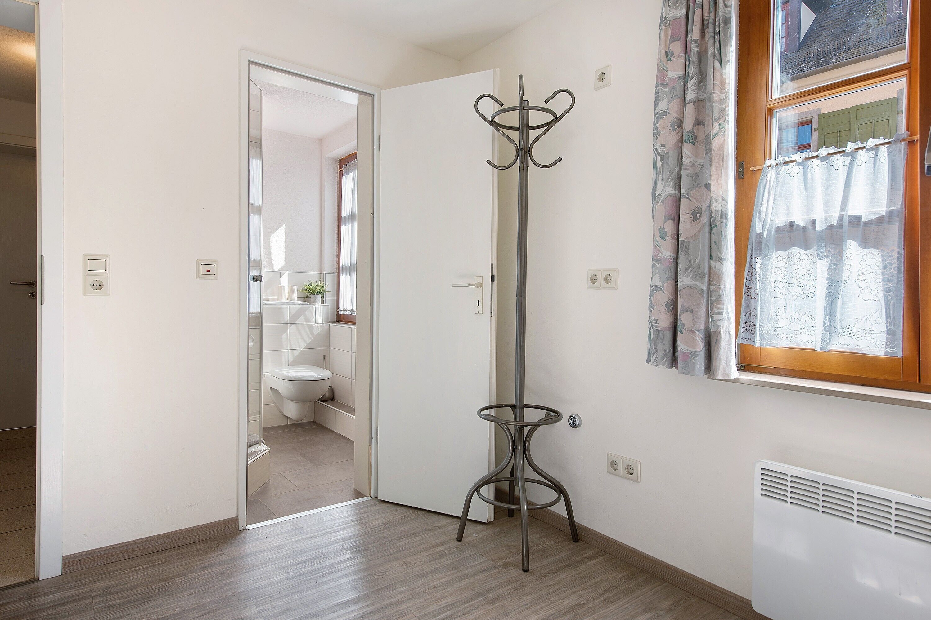 Une pièce avec un plancher en bois, un porte-manteau, une fenêtre avec des rideaux et une porte donnant sur une autre pièce.