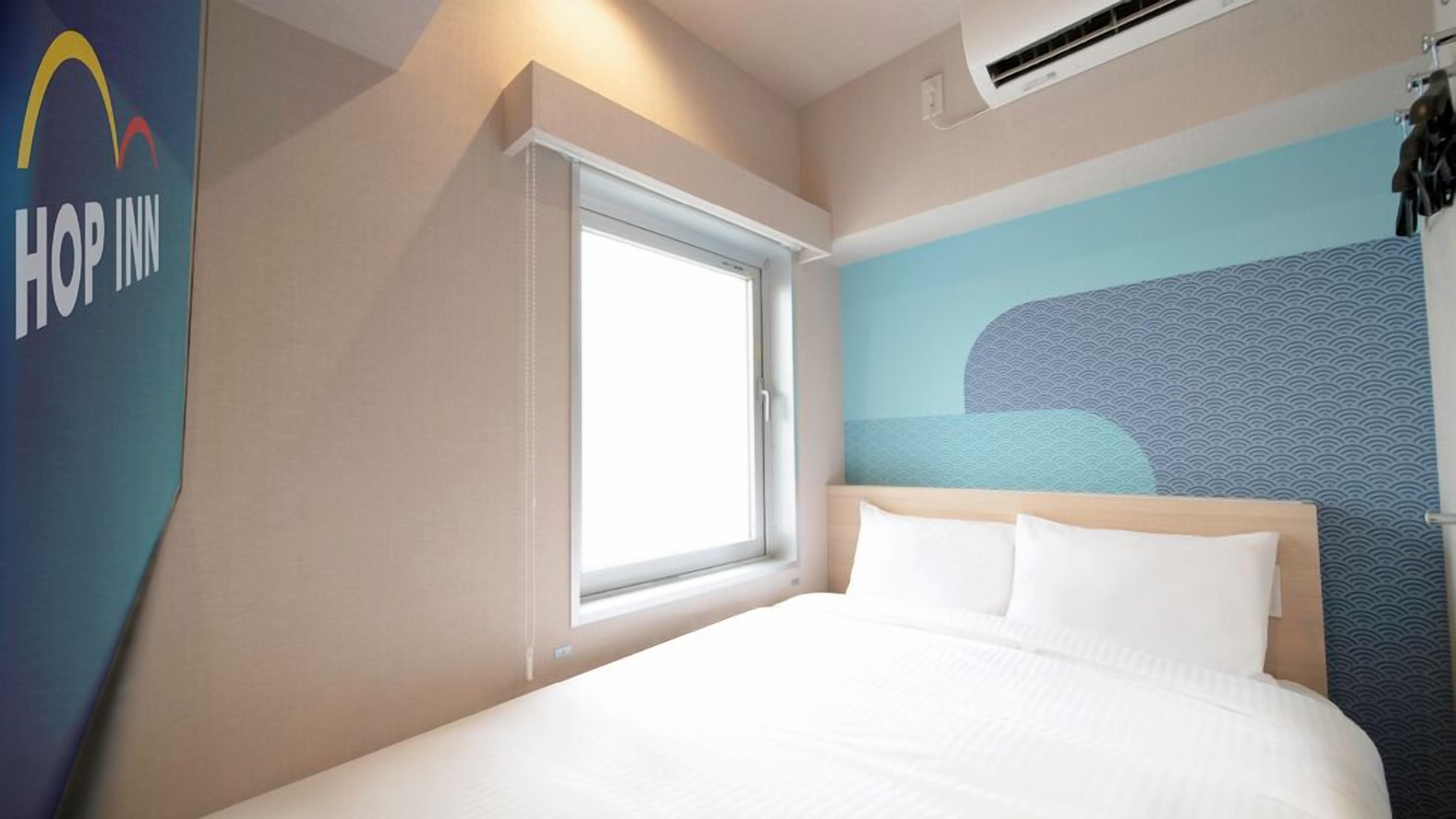 A hotel room with a bed, a window, a wall-mounted air conditioner, and a wall with a logo.