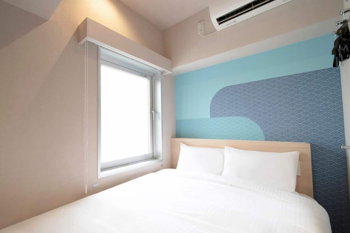 A hotel room with a bed, a window, a wall-mounted air conditioner, and a wall with a logo.