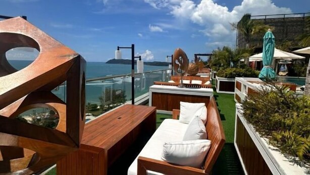 Un salón en la azotea con terraza de madera, asientos blancos acolchados y vista al océano.