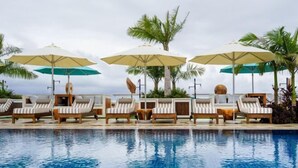 Un área junto a la piscina con sillones de descanso y sombrillas, rodeada de palmeras.