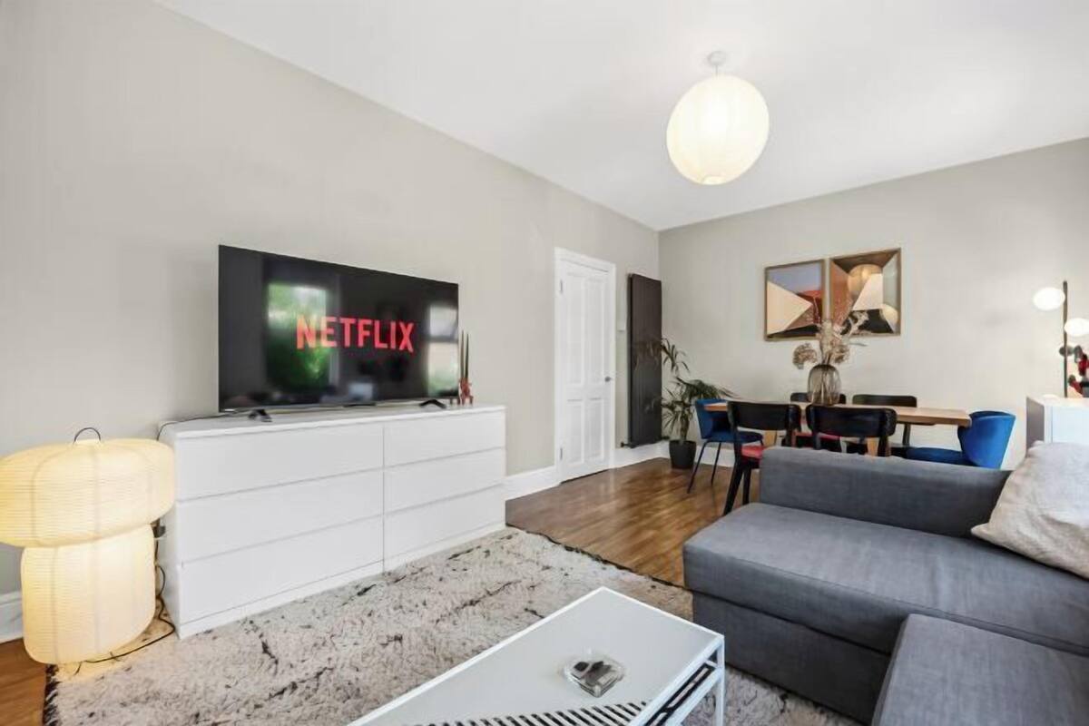 Un salon moderne avec un téléviseur à écran plat affichant le logo « NETFLIX », un canapé gris, un meuble blanc, une table basse et un coin repas avec des chaises et une table.