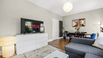 A modern living room with a flat-screen TV displaying 'NETFLIX', a grey sofa, a white cabinet, a coffee table, and a dining area with chairs and a table.