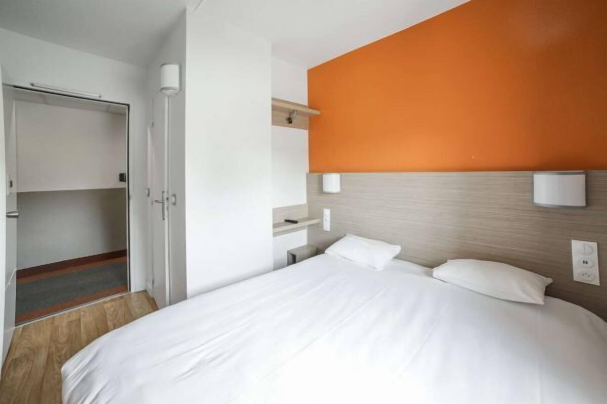 A hotel room with a bed, a wardrobe, a bedside table, and a wall-mounted light fixture.