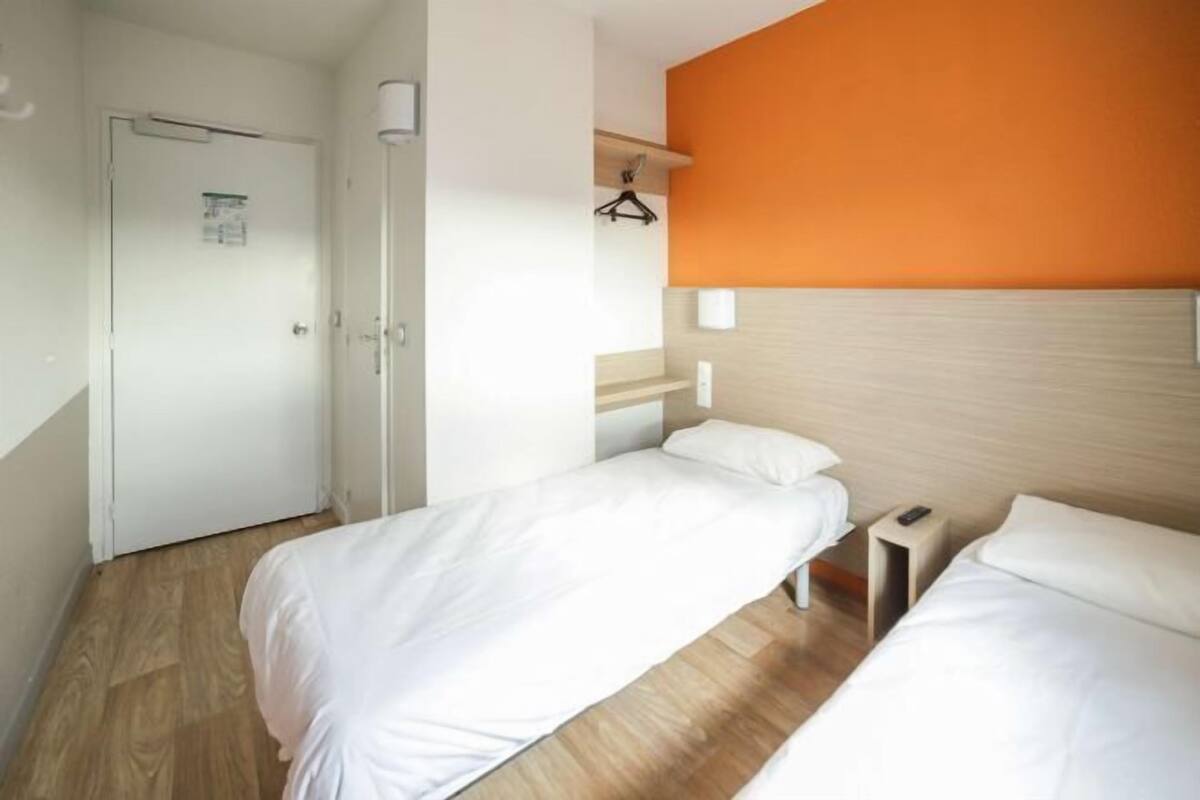 A hotel room with two single beds, a wooden floor, a white door, and a wall with an orange accent.