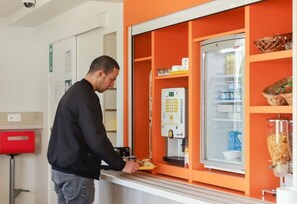 Un hombre preparando comida en una estación de autoservicio con una máquina de café y estantes refrigerados.
