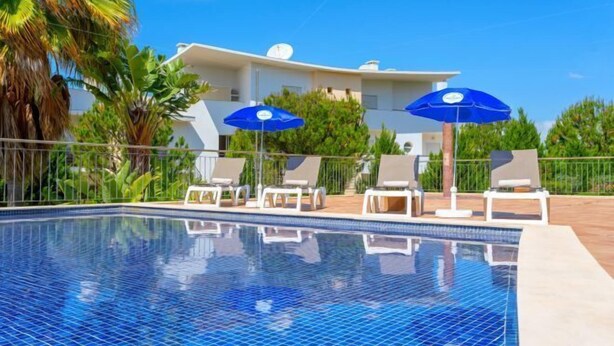 En swimmingpool med solstolar och parasoller, omgiven av palmer och en modern byggnad.