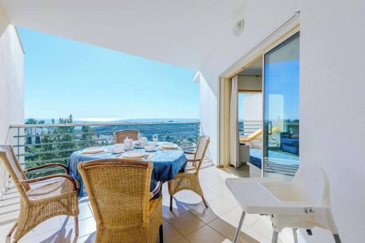 A balcony with a table set for two, wicker chairs, and a view of the sea.