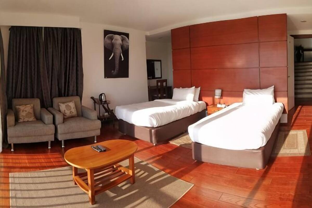 Une chambre d’hôtel avec deux lits, un canapé, une table basse en bois et un grand tableau représentant un éléphant accroché au mur.