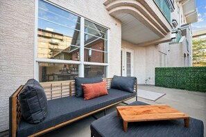 Uma área externa moderna com assentos, incluindo um sofá e uma mesa de madeira, janelas amplas e uma sebe verde.