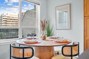 Uma sala de jantar com uma mesa redonda, cadeiras e vista para edifícios através de grandes janelas.