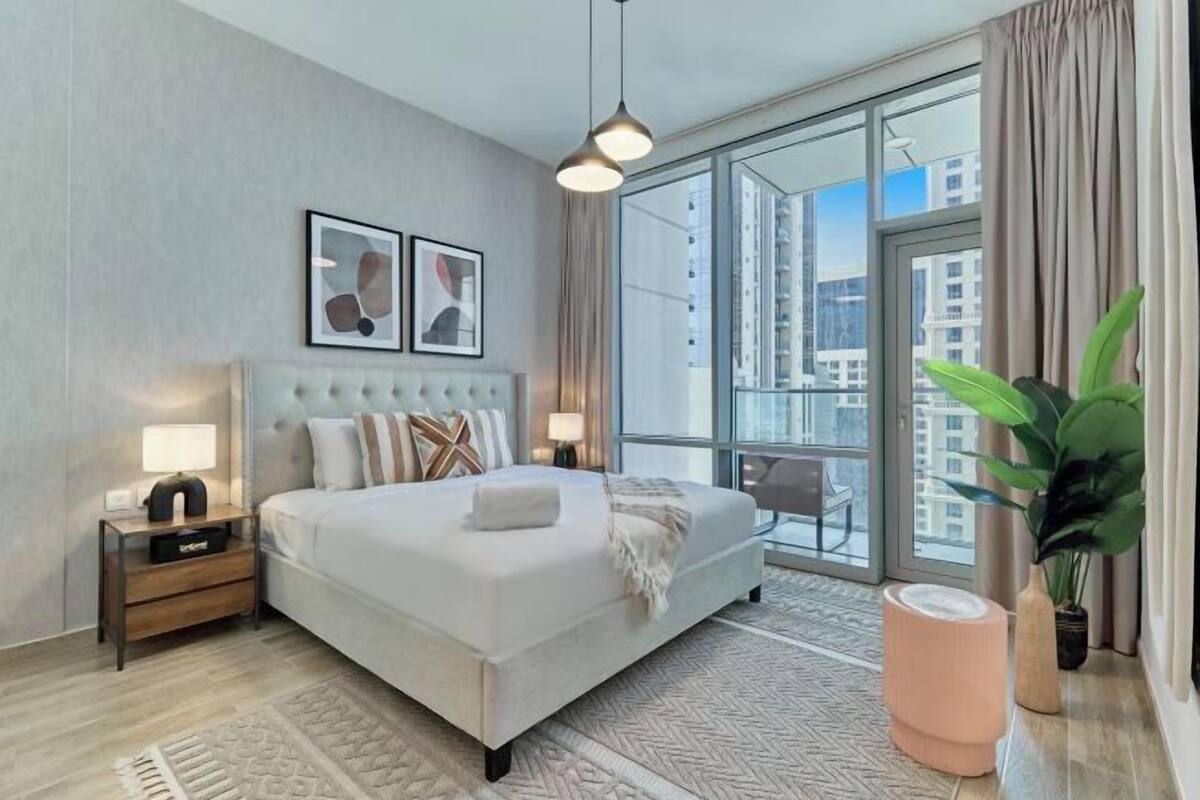 Une chambre d’hôtel moderne avec un grand lit, une table de chevet, une lampe, une grande fenêtre donnant sur des immeubles et une plante en pot.
