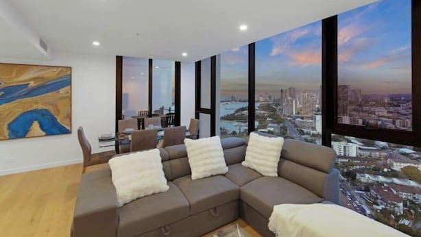 A modern living room with a sectional sofa, a painting on the wall, and large windows offering a city view. - Skyline Showdown - Views, Games & Vibes (Southport)