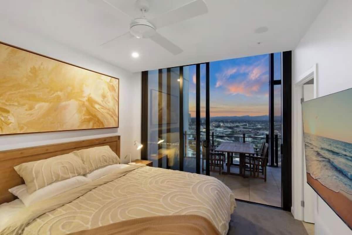 Une chambre avec un lit, une table de chevet, un ventilateur de plafond, une grande fenêtre donnant sur la ville et un tableau accroché au mur.