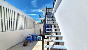 Un estrecho pasillo exterior con una escalera metálica que sube a un nivel superior. Hay taburetes de plástico azul y una mochila en el suelo.