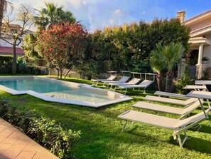 Una piscina con sillas de descanso, rodeada de follaje y un edificio.
