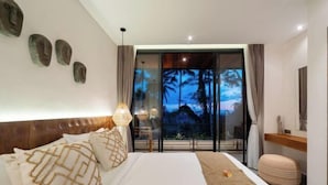 Una habitación de hotel moderna con una cama grande, vistas a palmeras a través de la puerta de vidrio, un cabecero de madera y una mesita de noche pequeña con una lámpara.