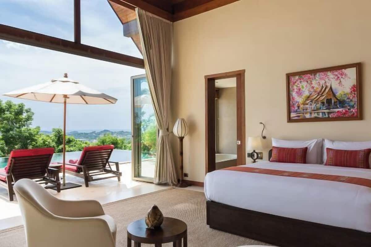 Ein Hotelzimmer mit einem großen Bett, einem Gemälde, Blick auf Grünflächen und einem Balkon mit Stühlen und Sonnenschirm.