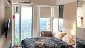 A modern hotel room with a large bed, a TV mounted on the wall, and a view of the city through the window. - PakuwonMall Apartment @Anderson Studio By B&B Host (Dukuhpakis)