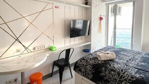 A modern hotel room with a bed, a desk with a chair, a TV, and a window with a balcony view. - PakuwonMall Apartment @Anderson Studio By B&B Host (Dukuhpakis)