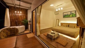 A modern bedroom with a large bed, a sitting area with a chair and table, and a view through a glass wall. - Merrin Farm (Mae Suai)