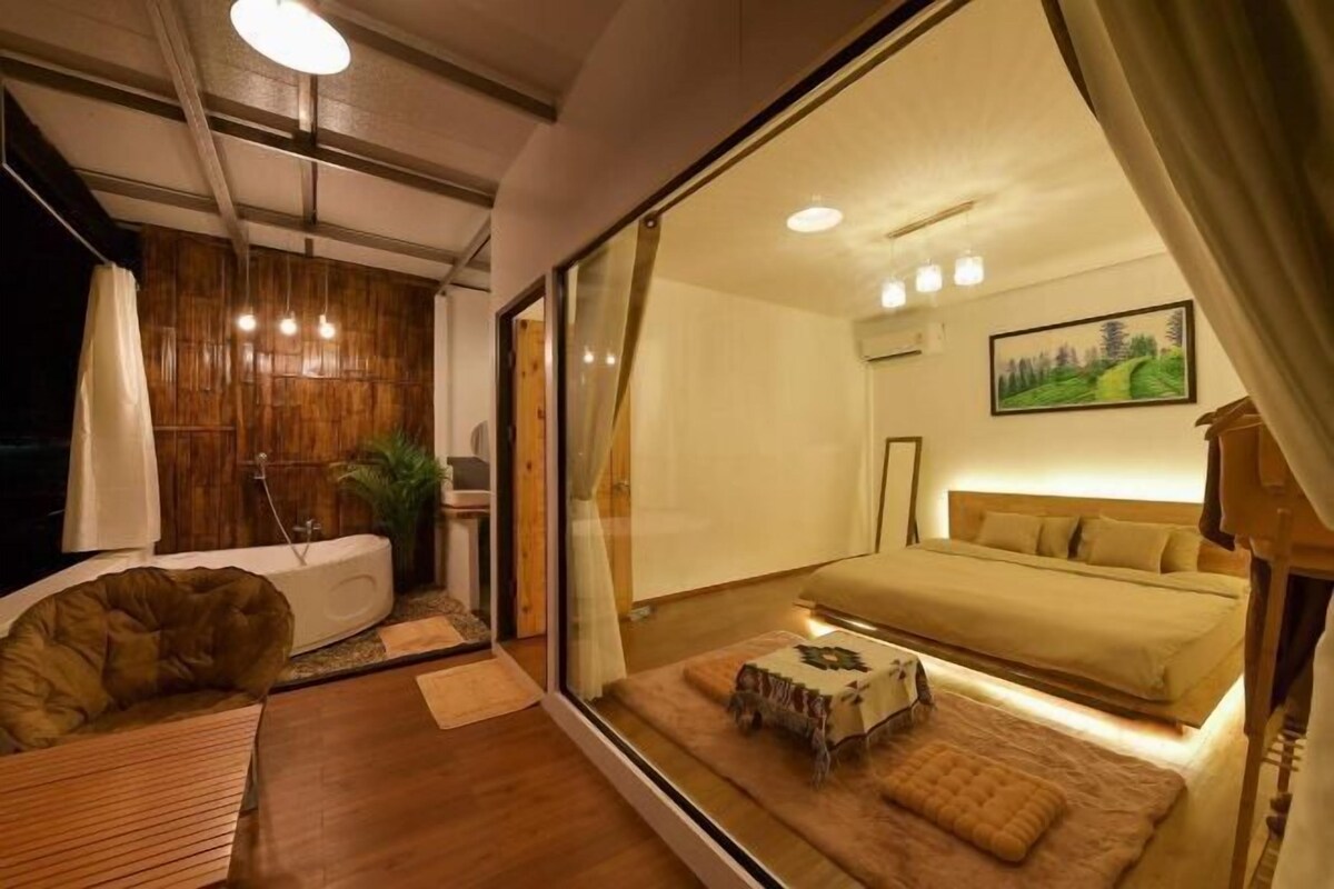 A modern bedroom with a large bed, a sitting area with a chair and table, and a view through a glass wall.