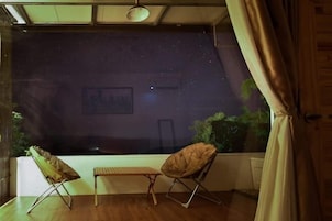 Una habitación con un ventanal grande, dos sillas plegables, una mesita y una cortina.