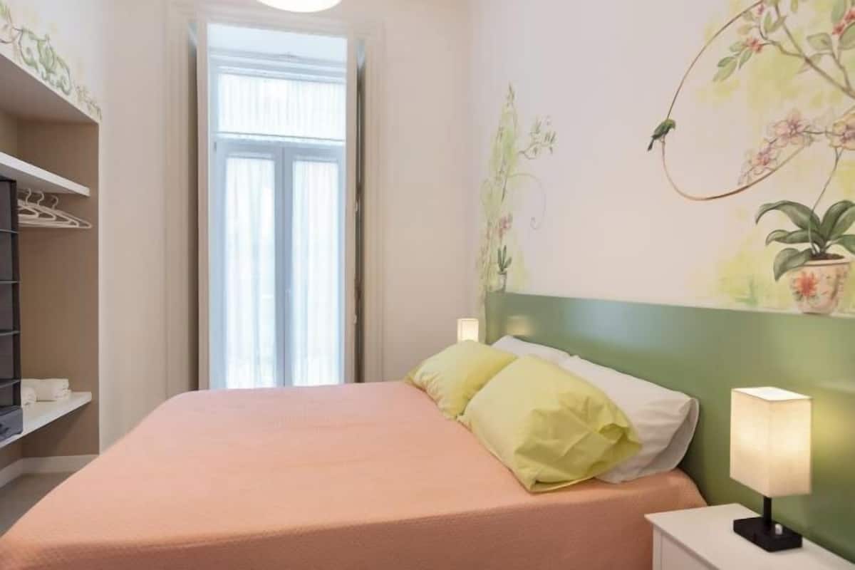 A neatly made bed with a peach-colored bedspread and two pillows, a bedside lamp, and a wall with a floral mural.