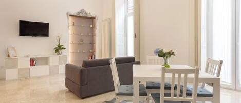 A modern living room with a sofa, dining table, and chairs, a flat-screen TV mounted on the wall, and a bookshelf.