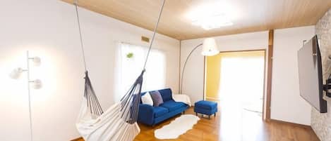 Sala de estar moderna com rede pendurada, sofá azul, tapete branco e chão de madeira.