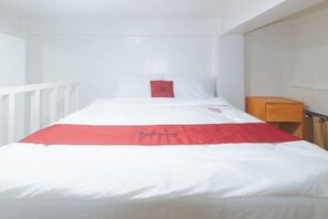 Een eenpersoonsbed met een wit-rode sprei, een rood kussen en een houten nachtkastje.